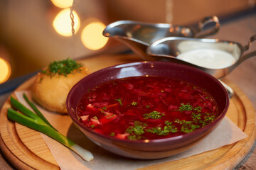A close photo of a Ukrainian borsch with a pampushka, green onion, and sour cream on a wooden tray on the table in a restaurant.