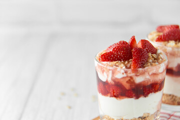 Homemade layered dessert with fresh strawberries, cream cheese or yogurt, granola and strawberry jam in glasses on white wood background. Healthy organic breakfast or snack concept. Selective focus.