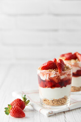 Homemade layered dessert with fresh strawberries, cream cheese or yogurt, granola and strawberry jam in glasses on white wood background. Healthy organic breakfast or snack concept. Selective focus.