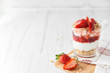 Homemade layered dessert with fresh strawberries, cream cheese or yogurt, granola and strawberry jam in glass on white wood background. Healthy organic breakfast or snack concept. Selective focus.