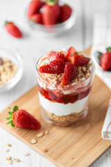Homemade layered dessert with fresh strawberries, cream cheese or yogurt, granola and strawberry jam in glass on white wood background. Healthy organic breakfast or snack concept. Selective focus.