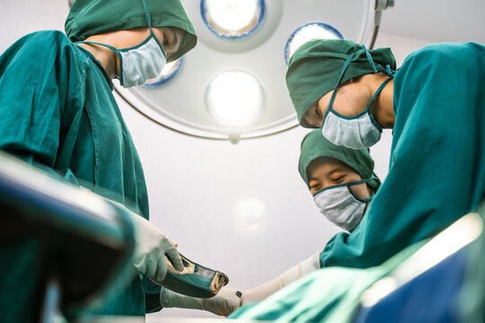 Professional medical team performing surgical operation in modern operating room. Asian assistant hands out instruments to surgeons doctor during operation on intensive care patient in hospital.