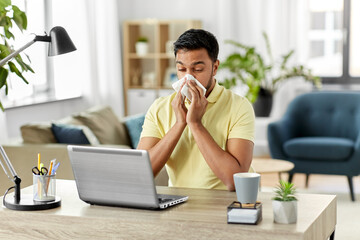 health, remote job and people concept - sick young indian man with laptop computer blowing nose at home office