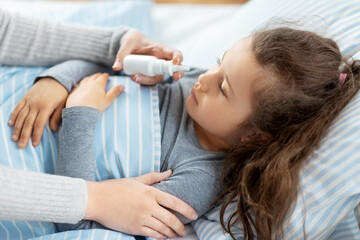 family, health and medicine concept - mother with nasal spray treats little sick daughter lying in bed at home