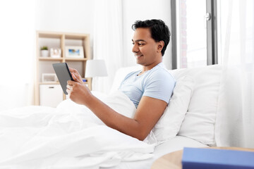 technology, bedtime and rest concept - happy smiling indian man with tablet pc computer lying in bed at home