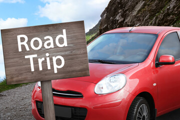 Wooden plaque with inscription road trip. Car parked near mountains on cloudy day