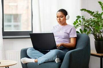 Naklejka premium people, technology and leisure concept - young african american woman in headphones with laptop computer listening to music at home