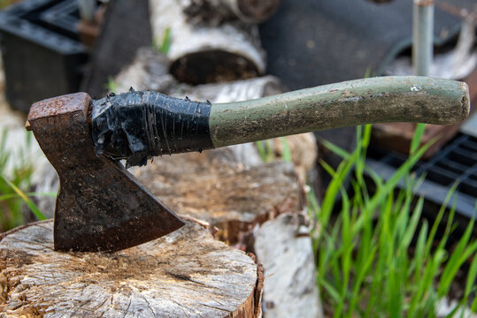 An Old Rusty Ax With A Wooden Handle Is Rewound With A Black Ribbon, Stuck In A Birch Stump Against A Background Of Green Grass And Birch Firewood. Chopping.