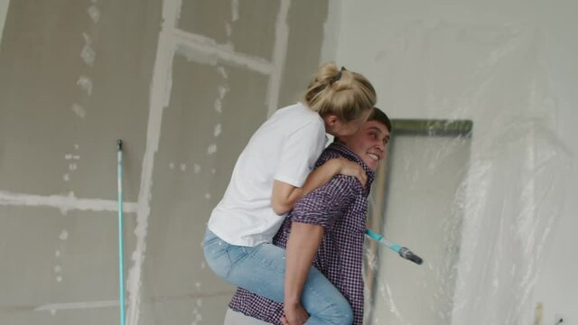 A happy girl looks at a guy who ride her on his back, a couple having fun while repairing an apartment