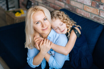 Happy woman and her curly little daughter are hugging at the home in cozy living room. Loving daughter gives bouquet flowers to her mother.Concept of Happy Mothers Day.