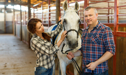 Smiling couple of farmers standing with white horse at stabling indoor