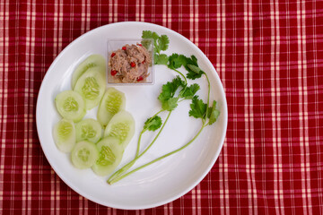 Yum small tuna salad in a clear plastic cup and place it on a white plate with coriander and sliced cucumber. Red plaid background. Top view.
