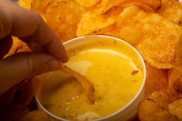 The girl takes a chip from a round dish with potato chips and a saucepan with cheese sauce in the center of the plate. Close up.