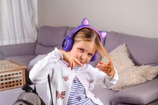Little girl in musical headphones and a backpack is ready to go to school. A European schoolgirl demonstrates positive emotions. Education and school days concept. Copy space