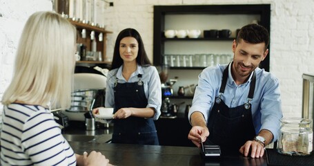 Back view on the blonde woman paying with a credit card to the waiter at the bar, while waitress serving her coffee.