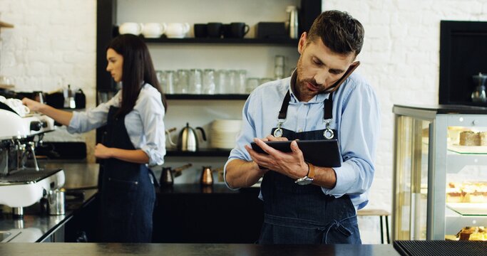 Caucasian attractive waiter speaking on the telephone and using tablet computer at the bar, brunette waitress making coffee behind.