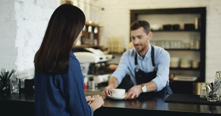 Handsome Caucasian barmen serving coffee to the pretty brunette woman at the bar, then she turning and smiling to the camera.