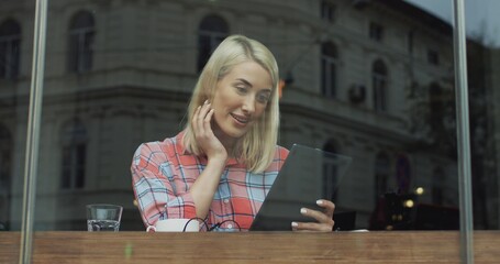 Attractive blond young woman sitting in the cafe with coffee, scrolling and taping on the tablet and watching something.