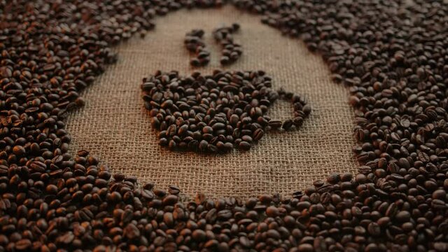 Figure of hot coffee mug with smoke assembled from natural arabica roasted beans. Coffee sign concept. Illustration in the centre of burlap cloth with scattered beans. Zooming in and focusing