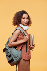 Portrait of positive pretty student girl with curly hair wearing satchel on back holding stack of workbooks against yellow background