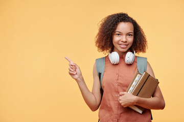 Portrait of smiling Afro-American student girl with books pointing aside while recommending online school