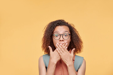 Emotional black student girl in eyeglasses shocked with news in education covering mouth with hands, yellow background