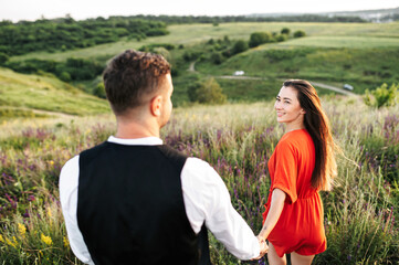 Back view of couple in love walking among tall grass in the meadow. Beautiful girl turned to the guy, a guy tenderly holds her hand