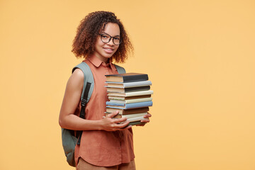 Portrait of smiling smart Afro-American student girl with satchel on back carrying stack of books against yellow background