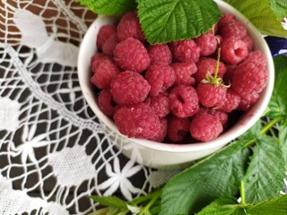 White ceramic bowl with ripe raspberries and green leaves on a lace tablecloth
