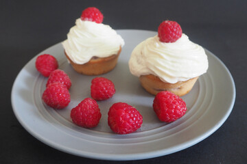 lots of raspberries on the background of two gluten-free cupcakes with cherries and mascarpone cream on a round gray plate . gluten-free homemade cakes
