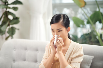 Sick young woman sitting on sofa blowing her nose at home in the sitting room. Photo of sneezing woman in paper tissue.