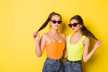 Two cheerful young hipster natural beauty girls posing together in sun glasses on yellow background