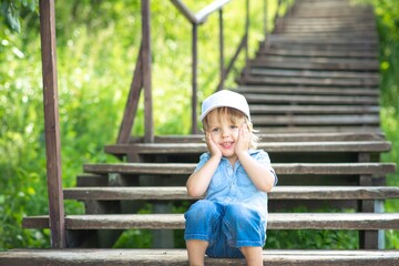 A boy in a white cap sits in a Park on a wooden staircase, posing and smiling.