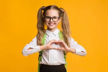 Fototapeta Smiling schoolgirl gesturing heart shape with fingers on yellow background obraz