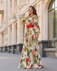 Fashion outdoor portrait. Elegant woman in colorful white red and green dress with floral design walking city street. Female model with long brunette wavy hair