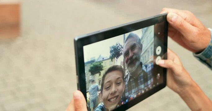 Close up of black tablet device in hands of teen boy and old man. Grandson and grandfather taking selfie photo on gadget camera outdoor at street. Screen of computer with photos of grandpa and son.