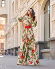 Fashion outdoor portrait. Elegant woman in colorful white red and green dress with floral design walking city street. Female model with long brunette wavy hair