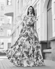 Fashion outdoor portrait. Elegant woman in colorful white red and green dress with floral design walking city street. Female model with long brunette wavy hair