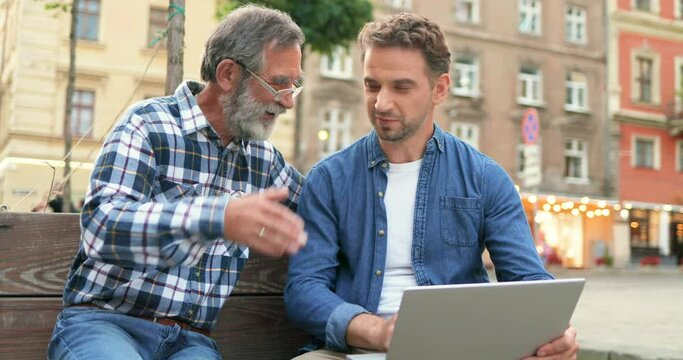 Handsome Caucasian adult son sitting at bench with old father and showing him something on computer outdoor at street. Two men of different ages and generations talking and using laptop.