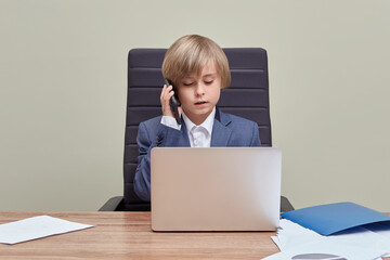 The blond boy works in the office with a laptop and talks on the phone.