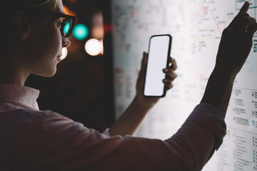 Interactive kiosk with public transport subway map.Female standing at big display with smartphone in hand.Young woman touching with finger screen while using train schedule application on mobile phone