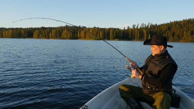 Fisherman In Black Cowboy Hat Fishing From Motorboat On Lake In Sunny Day With Reel Rod, Man Catching A Big Fish, Sport Fishing