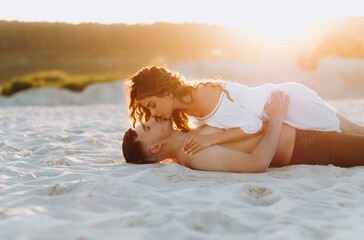 Incredibly beautiful couple lying on the sand at sunset. The girl in a white dress lies on the guy.