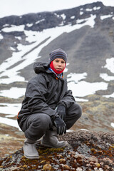 Fototapeta na wymiar Caucasian teenage boy dressing windproof and waterproof hiking outfit for mountaineering, young man sitting on rock and looking at camera in mountains