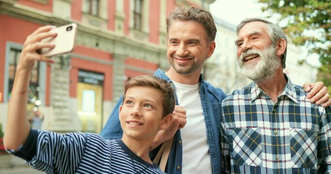 Cute teen Caucasian boy taking selfie photo with father and senior grandfather at street with smartphone. Old man with beard with adult son and small grandson posing while making photos on phone.