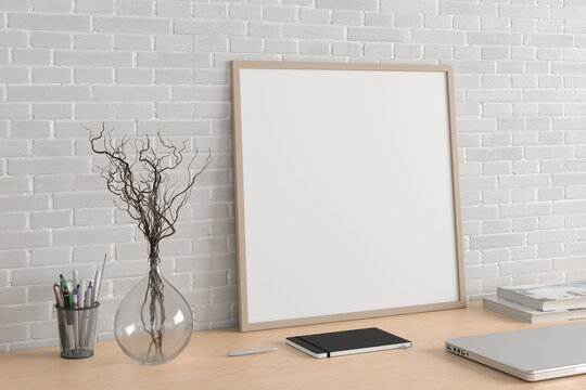 Square poster frame mockup on the wooden table of home studio workspace with white brick wall. Side view, clipping path around poster picture.