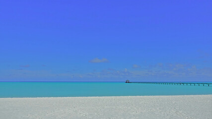Fototapeta na wymiar Maldivian minimalism. White sand beach, aquamarine ocean, azure sky. Above the water there is a wooden footpath and a canopy in the distance. 