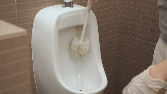 Women hand in gloves cleaning urinal bowl with brush.