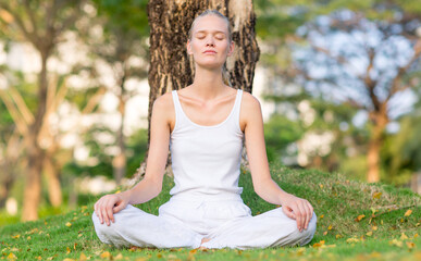 Serene young woman meditating outdoor at peace on the grass under a tree at the park on a calm morning. Self care, mind and body relaxation.