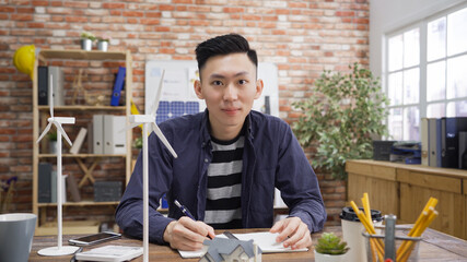 Confident asian business man looking at camera smiling with models of windmills turbines on wood table. young guy architect working with alternative energy construction project indoor modern office.